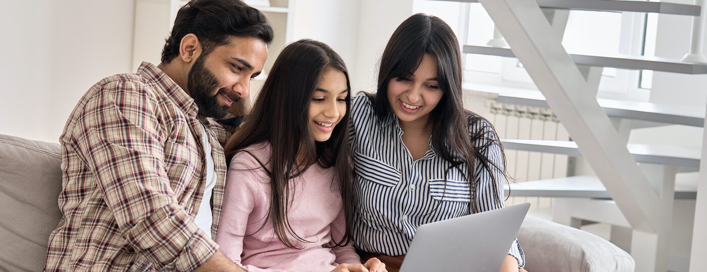 Happy indian family couple with child daughter using laptop computer at home. Smiling parents and teen kid bonding watching streaming online tv or doing ecommerce shopping together sitting on sofa. Happy indian family couple with child daughter using laptop computer at home. Smiling parents and teen kid bonding watching streaming online tv or doing ecommerce shopping together sitting on sofa.
