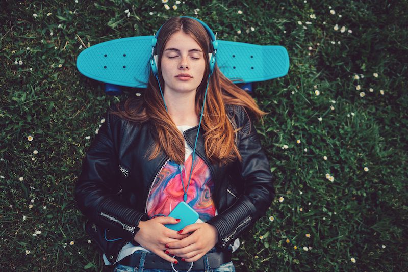 Relaxed teenage girl in the grass listening to music Young woman lying down in the grass with skateboard behind head