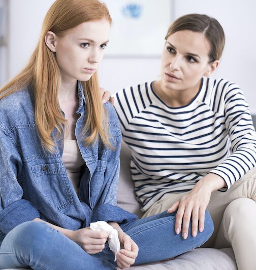 Woman comforting an emotional young teenage girl sitting with her legs crossed on a sofa and crying