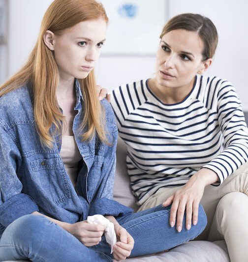 Woman comforting emotional teenage girl Woman comforting an emotional young teenage girl sitting with her legs crossed on a sofa and crying