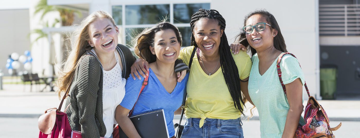 A group of four multi-ethnic teenage girls, 18 years old, standing in front of a school building carrying book bags and notebooks. They are high school seniors or freshmen in college.